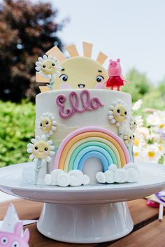 a birthday cake decorated with flowers and rainbows on a table in front of trees