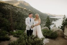 a bride and groom standing on top of a mountain next to each other holding hands