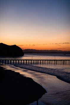 the sun is setting over the ocean with a long pier in the foreground and mountains in the distance