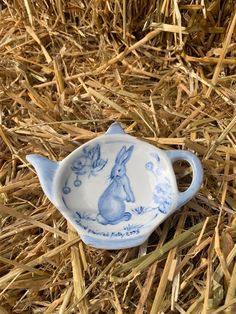 a blue and white porcelain tea pot sitting on straw in the grass with an image of a rabbit painted on it's side