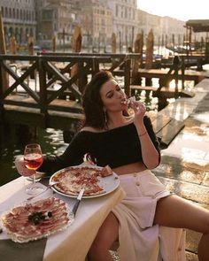 a woman sitting at a table eating pizza and drinking wine in front of the water