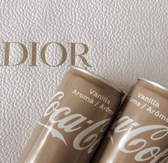 two cans of coca - cola sitting on top of a white surface with the word dior written in gold