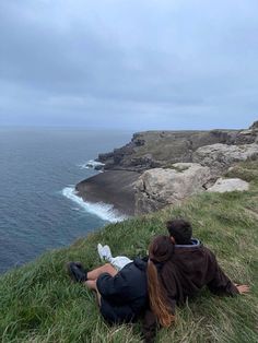 two people are sitting on the grass by the water looking out at the ocean and cliffs