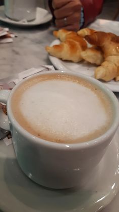 a cup of cappuccino on a plate with croissants in the background