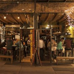 people sitting at tables in front of a bar with lights on the ceiling and windows