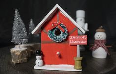 a small red birdhouse with a wreath on it's door and two snowmen next to it