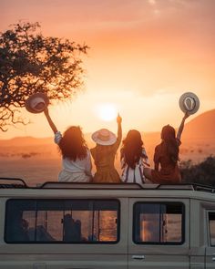 three women sitting on the roof of a vehicle with their arms in the air as the sun sets