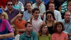 a group of people sitting next to each other at a tennis match with one man holding up his hand