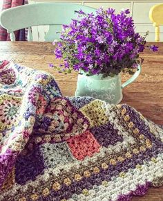 a crocheted blanket sitting on top of a wooden table next to a vase filled with purple flowers