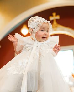 a baby dressed in a white dress and bonnet is standing on the floor with her arms outstretched