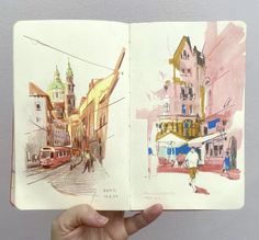 a hand holding up an open book with watercolor drawings on the pages and buildings in the background