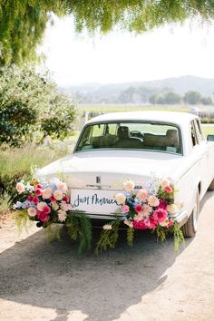 an old white car with flowers on the front is parked under a tree and has a sign that says, our mama