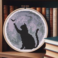 a cross - stitch picture of a cat on a shelf with books in the background