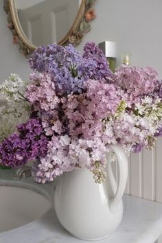 purple flowers are in a white vase on a table next to a mirror and dresser