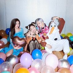 three women sitting in a room with balloons all around them and one woman holding a stuffed animal