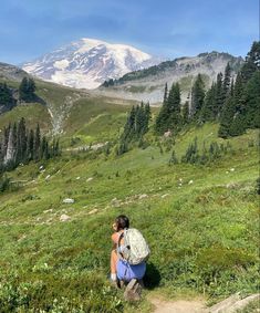 a woman kneeling down on top of a lush green hillside next to a snow covered mountain