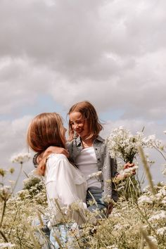 two young women standing in a field of tall grass and wildflowers looking at each other