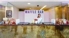 a woman standing behind a counter in front of a sign that says waffle bar