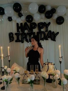 a woman standing in front of a table filled with cake and candles, surrounded by black and white balloons