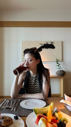 a woman sitting at a table drinking from a bottle with fruit on the plate next to her