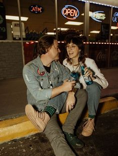 a man and woman are sitting on the curb with sodas in their hands as they look at each other