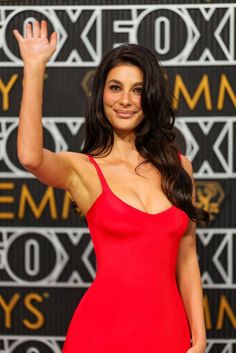 a woman in a red dress waves at the camera while standing on a red carpet