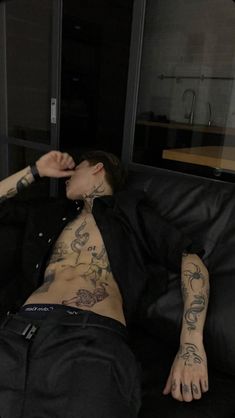 a man with tattoos on his chest laying on a black leather couch in front of a window