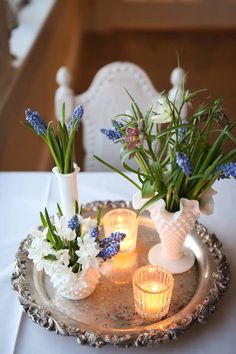 vases with flowers and candles are on a silver tray in front of a white chair