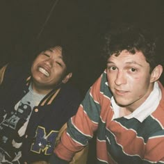 two young men sitting next to each other in front of a black background, one smiling and the other looking at the camera