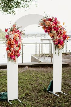 two white archways decorated with flowers and greenery on the grass near water in front of a pier