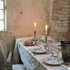 a table with plates and candles on it in front of a brick wall, next to a window