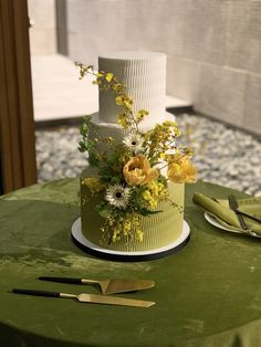 a three tiered cake with yellow flowers and greenery sits on a green tablecloth