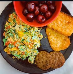 a black plate topped with fried eggs, meat and vegetables next to a bowl of fruit