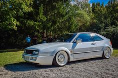 a silver car parked on top of a gravel road next to trees and people standing around