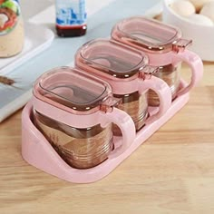 three pink glass containers with lids on a wooden table next to eggs and other ingredients