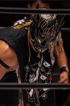 a man wearing a black mask with tattoos on his arm and chest, standing next to a metal fence