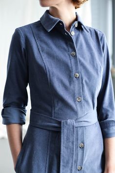 a woman wearing a blue shirt dress with buttons on the front and side, standing next to a window