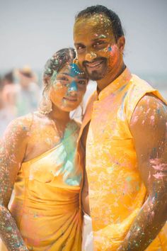 a man and woman standing next to each other covered in colored powder on the beach