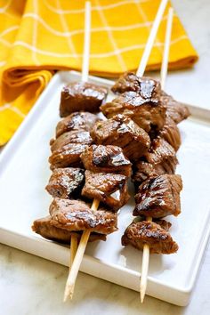 several skewers of meat sitting on a white plate next to a yellow napkin