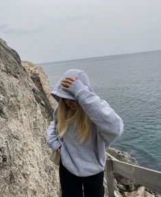 a woman standing on the edge of a cliff next to the ocean wearing a hoodie