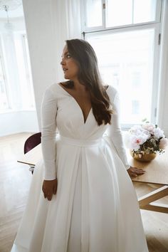 a woman standing in front of a window wearing a white wedding dress with long sleeves