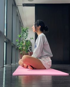 a woman sitting on a pink yoga mat in front of a window with a potted plant