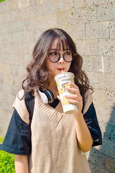 a woman wearing headphones and holding a drink in front of a brick wall with grass behind her