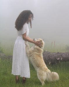 a woman in white dress petting a large dog on the side of a foggy field