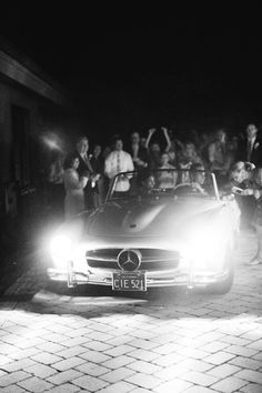 a black and white photo of a car at night with people in the background watching