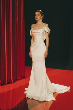 a woman standing in front of a red curtain wearing a white dress with one shoulder