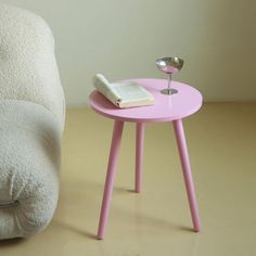 a small pink table with a book on it next to a white chair and lamp