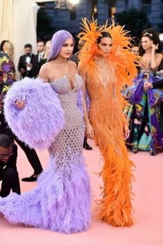 two women dressed in purple and orange feather gowns pose for the camera while others look on