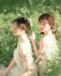 two young women sitting in the grass with flowers on their heads and one wearing a white dress