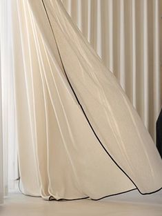 a woman in a long white dress standing next to a window with curtains on it